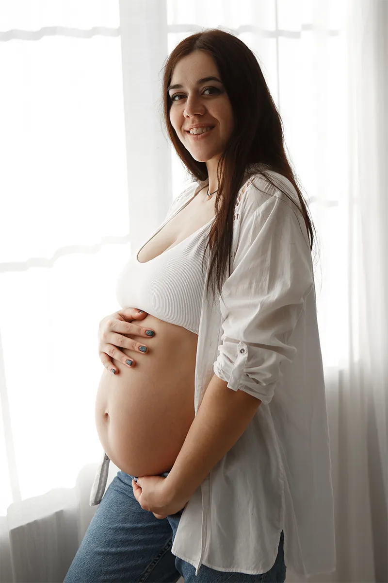 Gestante de camisa branca com as mãos sobre a barriga, perto de uma janela
